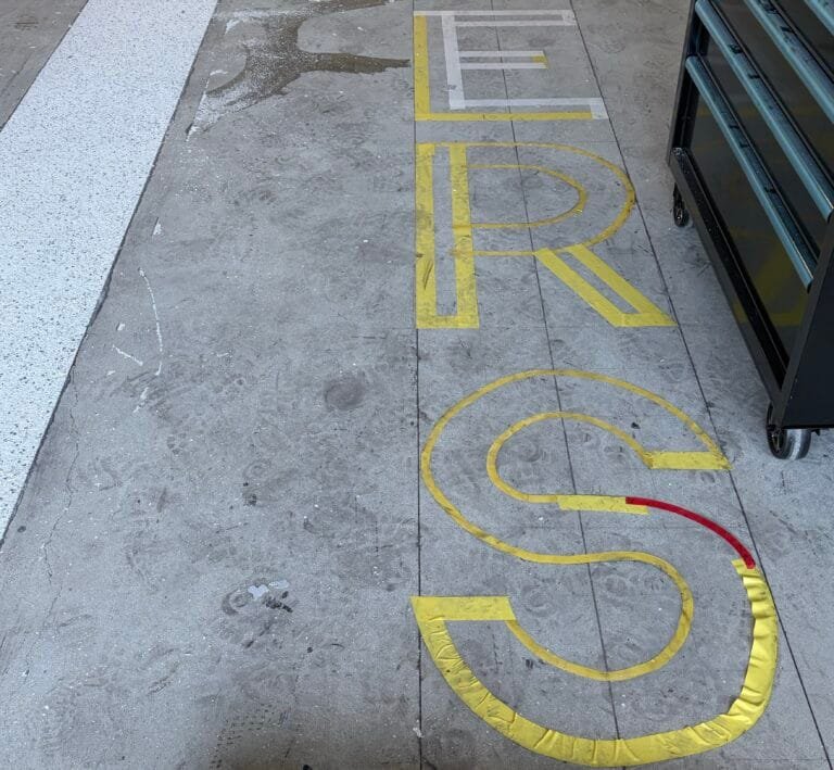 Prepared garage slab with the word HERS cut and masked in yellow tape, ready for a second or third flake colour.